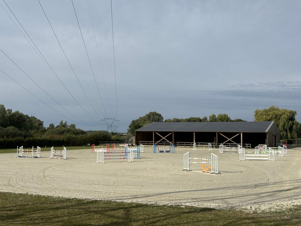 Carrière extérieure équipée d'obstacles colorés devant le hangar de l'écurie.