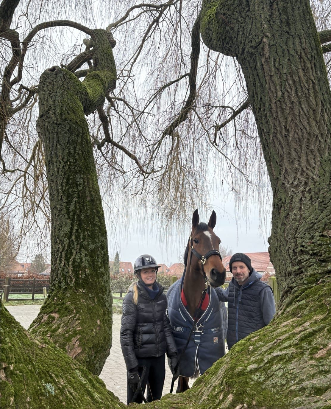 Deux membres de l'équipe posant avec un cheval bai entre deux grands saules.
